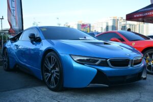 A sleek blue BMW i8 sports car is parked in an outdoor area of Las Vegas, showcasing its aerodynamic design and modern styling. It sits on stylish alloy wheels. To its right, part of a red sports car and a red canopy tent are visible. Buildings in the background are set against a clear sky.